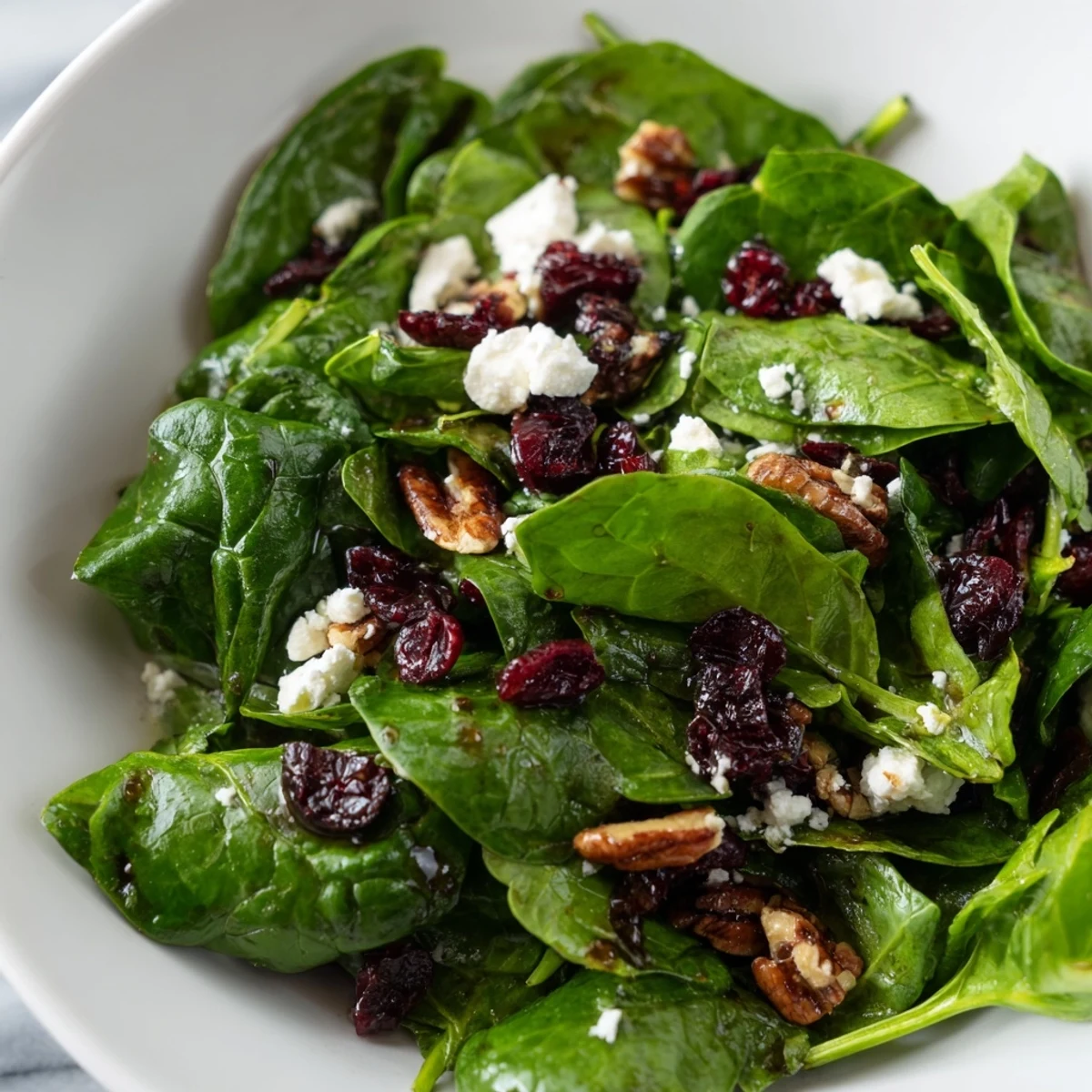 A close-up of a Spinach Goat Cheese Salad, showing glossy balsamic drizzle over tender greens, candied pecans, and soft crumbles of goat cheese.