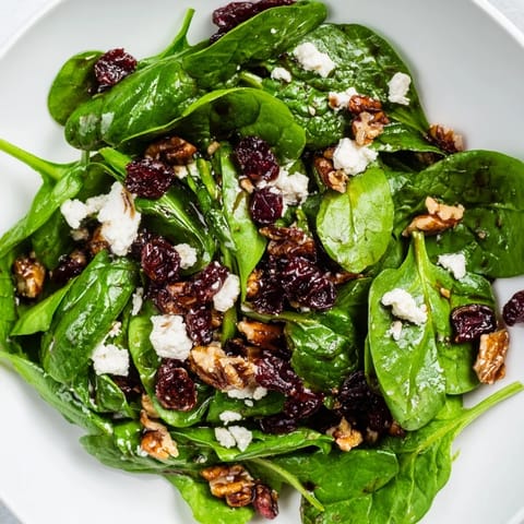 Image of a fresh Spinach Goat Cheese Salad with crumbled goat cheese, sweet cranberries, and crunchy candied pecans on a textured white plate.